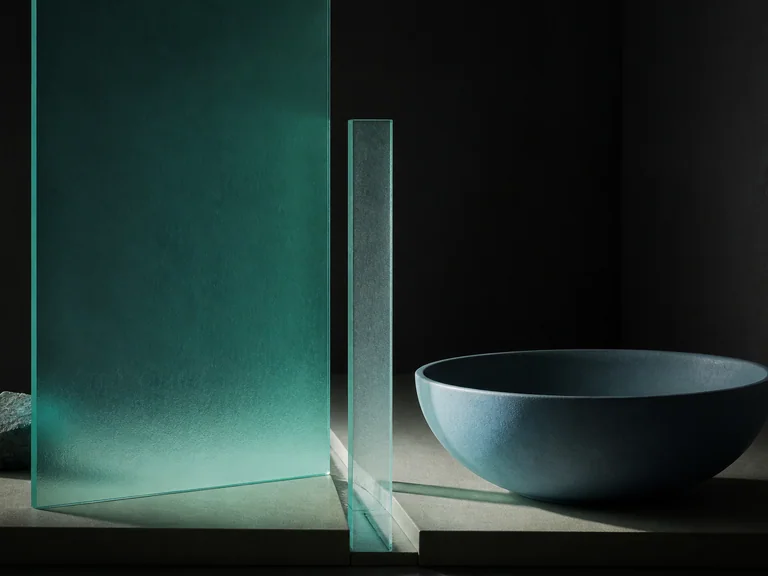 A tall teal glass plane and narrow translucent strip face a low blue basin across an uneven channel.